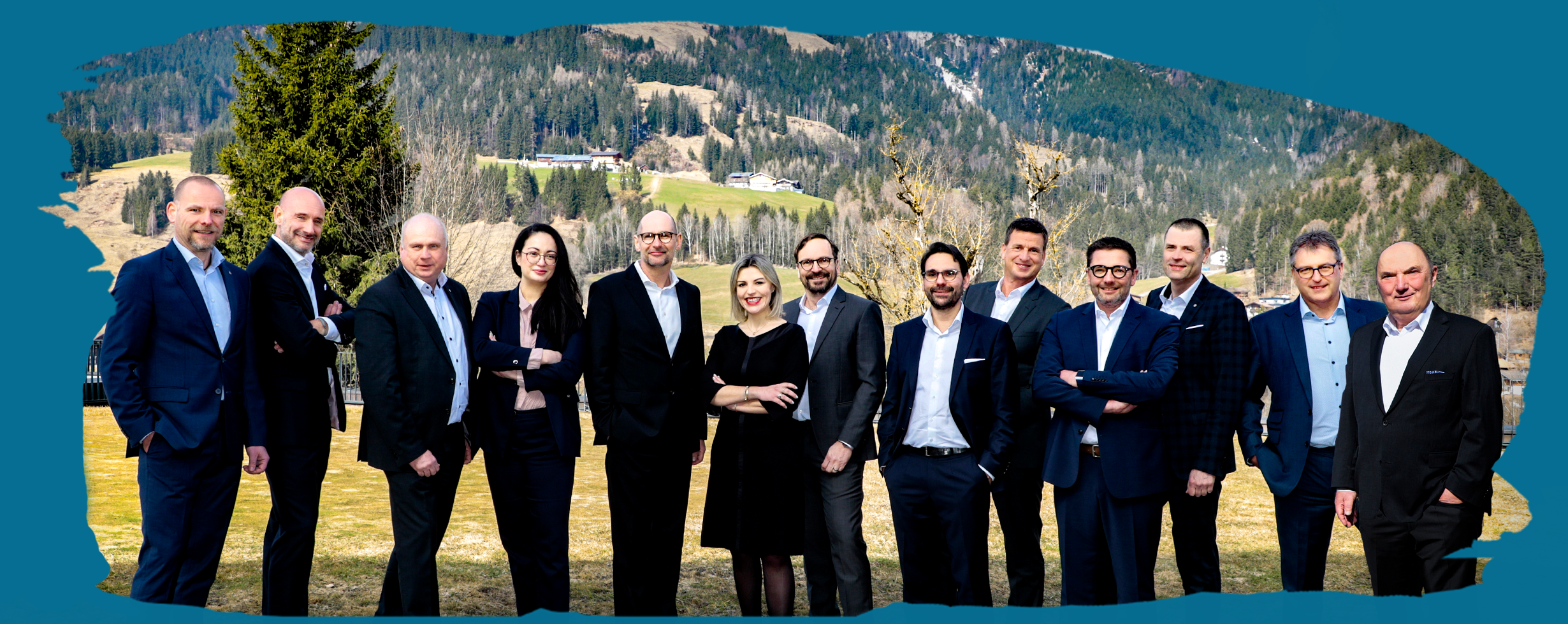 Man sieht die 13 leitenden Personen der Wackler Group nebeneinander auf einer Wiese stehend. Sie tragen Anzug, Blazer oder Kleid. Im Hintergrund ist eine Berglandschaft zu erkennen.